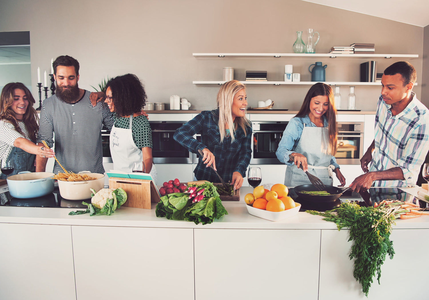 A group of six diverse friends, Black and Caucasian, gathered in a kitchen cooking and enjoying food together, with sharpened knives on the counter.