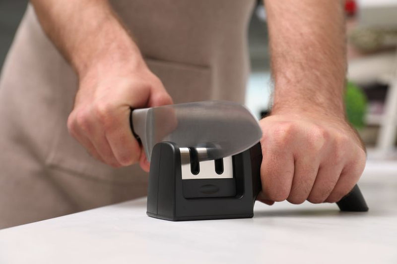 Man sharpening knife with pull-through sharpener at white table indoors, closeup