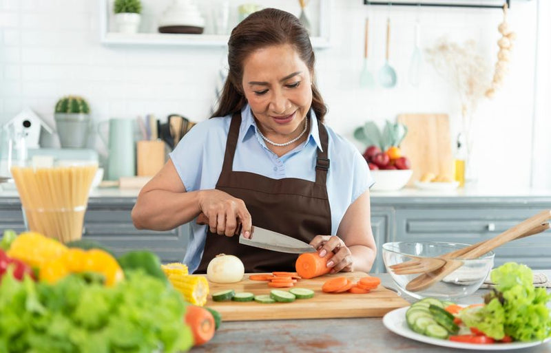 Happy smiling Asian older woman in apron cutting vegetables with sharp kitchen knife