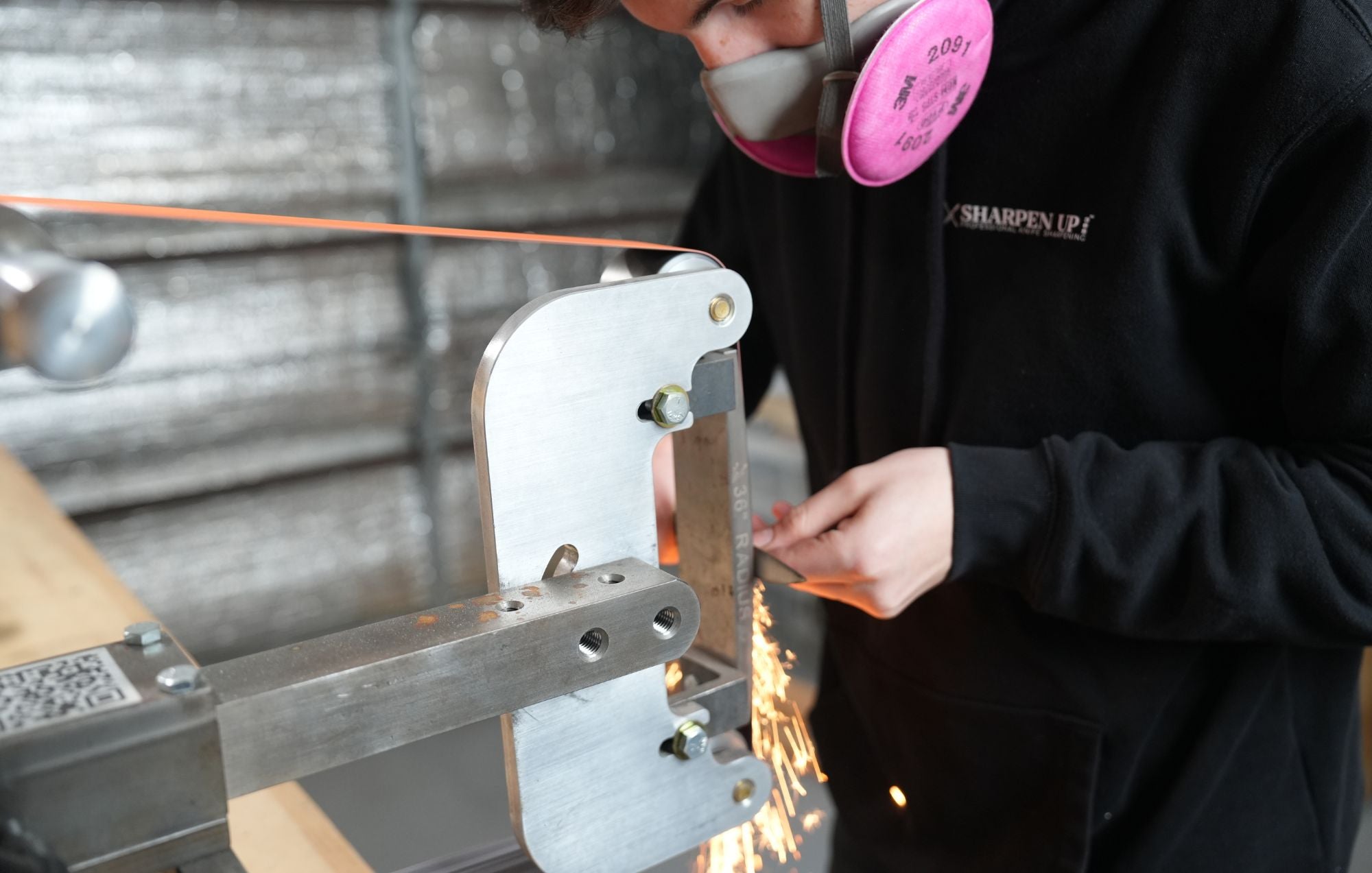 Person sharpening a knife with sparks flying, wearing a black shirt and pink respirator.